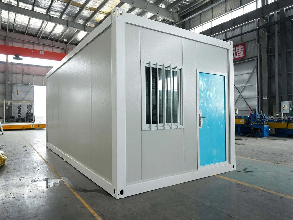 A white modular container home with a bright blue door and a barred window, photographed inside a large industrial warehouse.