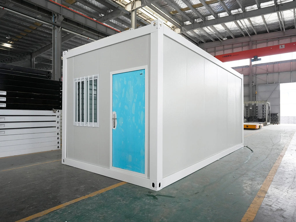 A white modular shipping container home with a bright blue door and a small barred window, photographed inside a large industrial warehouse.