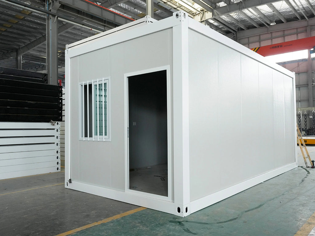 A white modular shipping container turned into a small office or cabin, shown in a warehouse setting with its front door open.