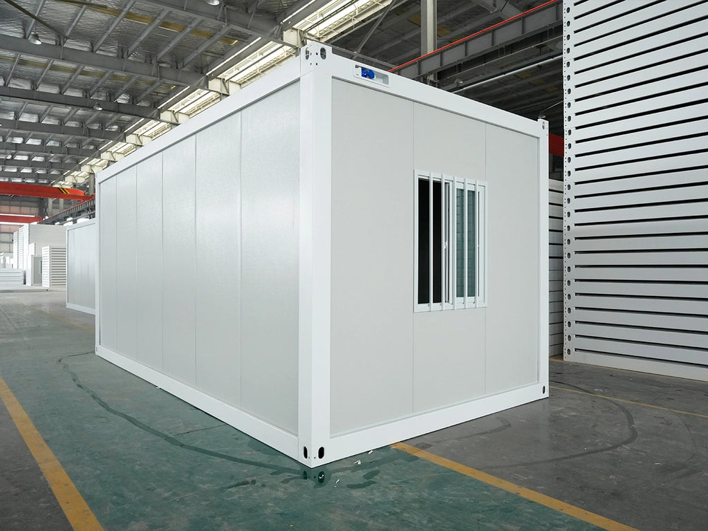 A white modular container office or living space with a small window, photographed inside a large warehouse or factory interior.