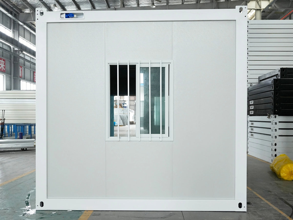 A large white modular office or utility container with a small barred window, photographed in a warehouse or factory setting.