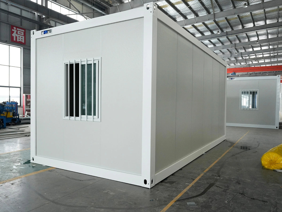 A white modular container office or living space with a barred window, photographed inside a large warehouse or factory. Another similar container is visible in the background.