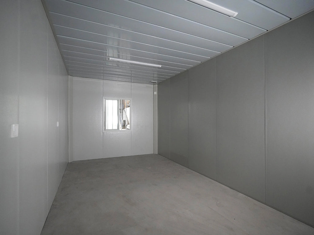 An empty industrial room with gray panel walls, a gray concrete floor, a white paneled ceiling with fluorescent lights, and a small window at the far end.