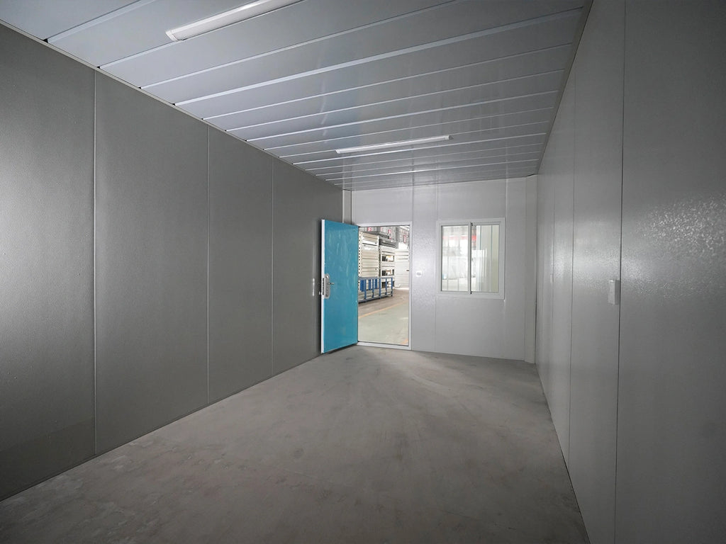 A long, narrow industrial room with gray panel walls, a gray concrete floor, a white ceiling with fluorescent lights, and an open bright blue door at the far end revealing another interior space.