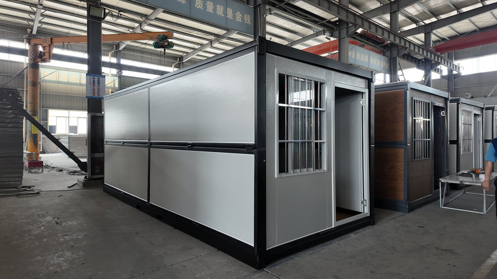 A modular container-style office or living unit with white exterior panels and a black frame, shown inside a large warehouse or factory. The main unit has a barred window, an open doorway, and a small table placed outside it.
