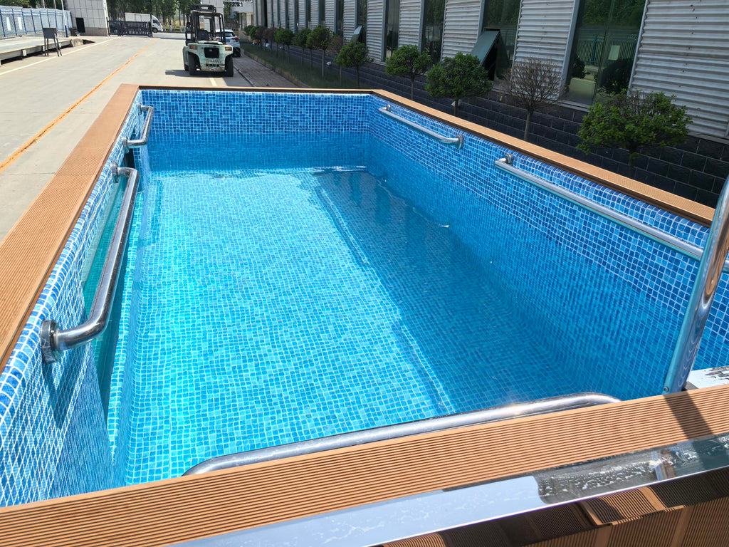 Rectangular pool with blue tiles and wooden deck area, surrounded by a building and vehicles.