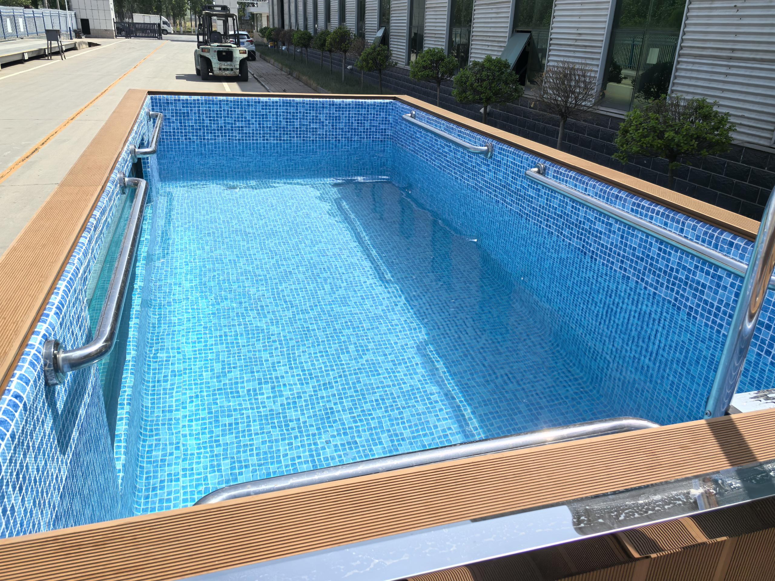Rectangular pool with blue tiles and wooden deck area, surrounded by a building and vehicles.