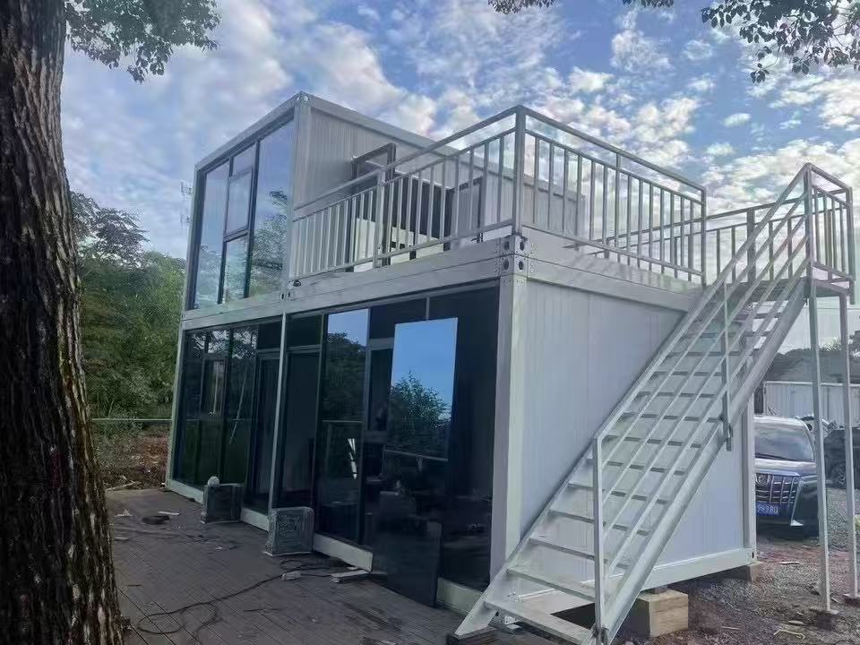 Two-story container house with a staircase and deck, surrounded by trees and a clear sky.